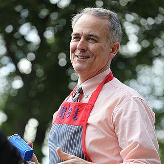 Photo of John McVay smiling wearing a striped apron and holding a camera and blue cup